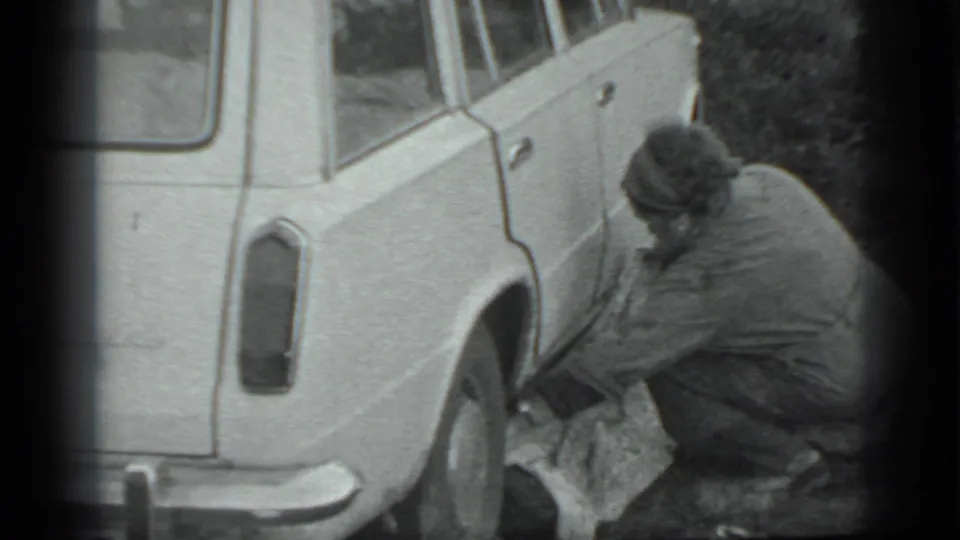 The man remains crouched beside the station wagon in Moscow, handling a cloth-covered item near the rear wheel.