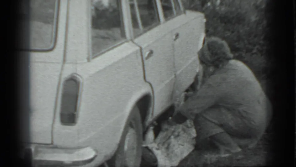 A dark-coated man crouches beside the white station wagon, partly turned away as he works near the rear wheel.