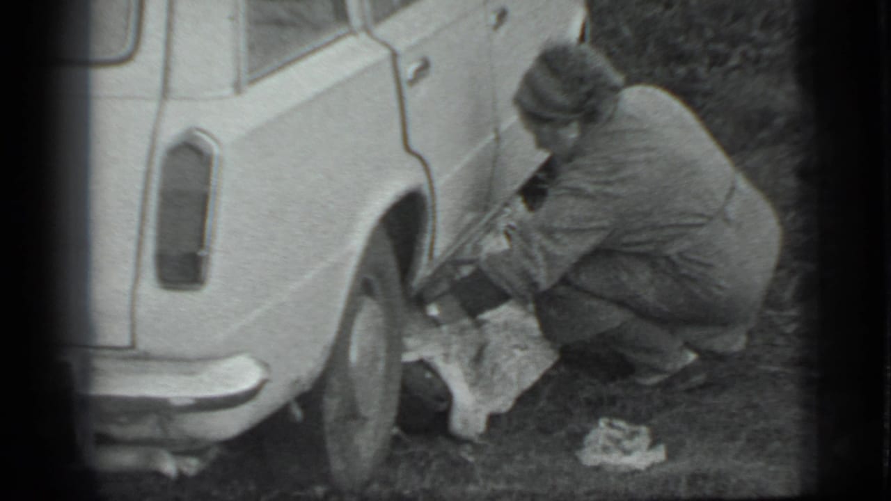In Moscow 1983 footage, a woman in a coat and headscarf crouches by the rear of a light-colored car near a cloth on the ground.