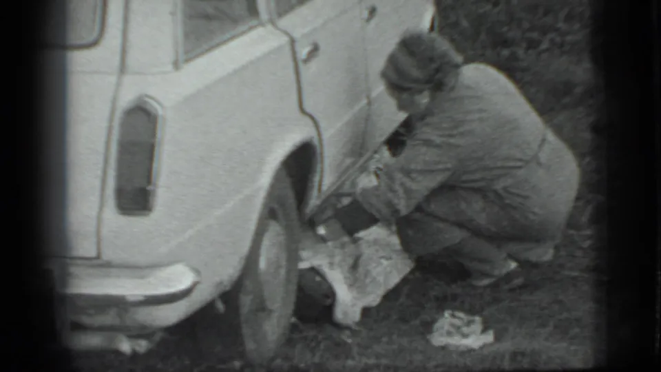 In Moscow 1983 footage, a woman in a coat and headscarf crouches by the rear of a light-colored car near a cloth on the ground.