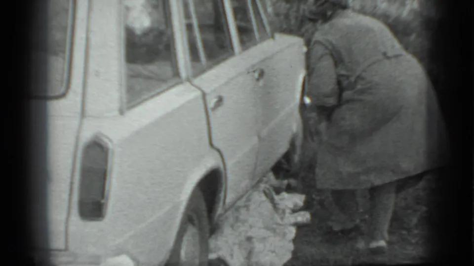 The woman bends near the side of the light-colored car, appearing to adjust or cover something with a cloth beside the vehicle.