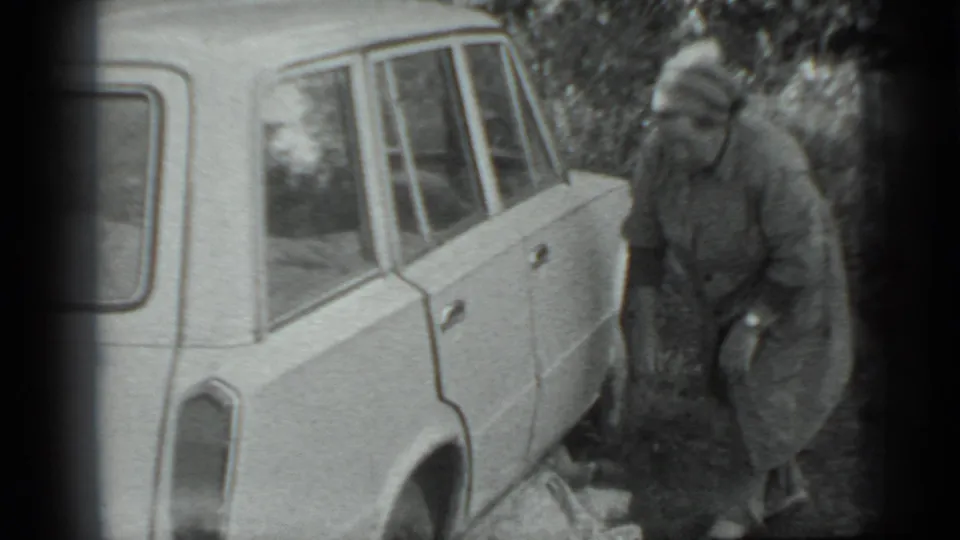 A woman in a long coat and headscarf stands beside the light-colored car, with a cloth or blanket visible near her feet.