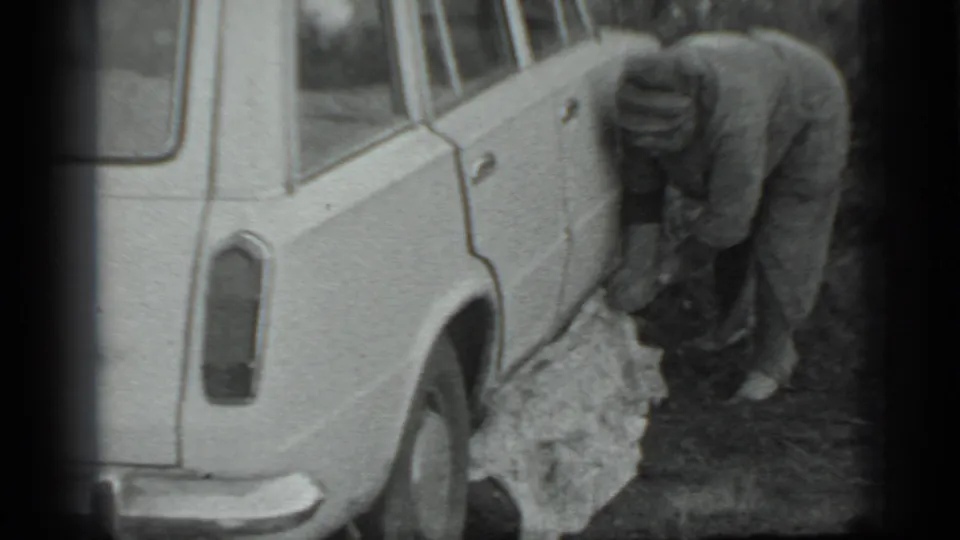The woman bends again near the rear side of the car, handling a light cloth or blanket on the ground in the 1983 Moscow home-movie scene.