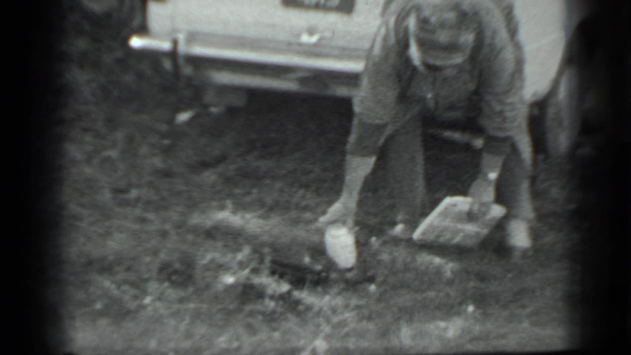 In metadata-supported 1983 Moscow footage, a woman bends beside a light-colored car to gather white objects from the grass.