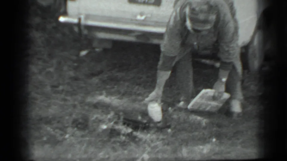 In metadata-supported 1983 Moscow footage, a woman bends beside a light-colored car to gather white objects from the grass.
