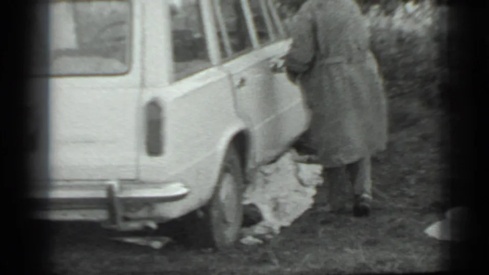 In the final frame, the woman turns away from the car with pale items in hand, seen in grainy 8mm outdoor footage.