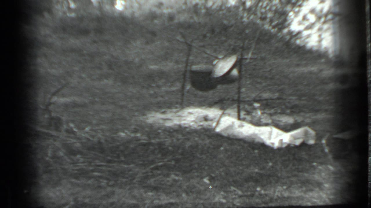 Black-and-white 1983 Moscow home-movie frame showing a pot suspended from tripod poles at a campsite, with cloth or tarp on the ground.
