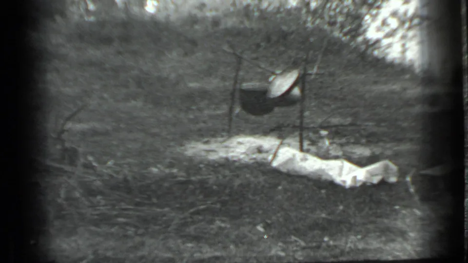 Black-and-white 1983 Moscow home-movie frame showing a pot suspended from tripod poles at a campsite, with cloth or tarp on the ground.