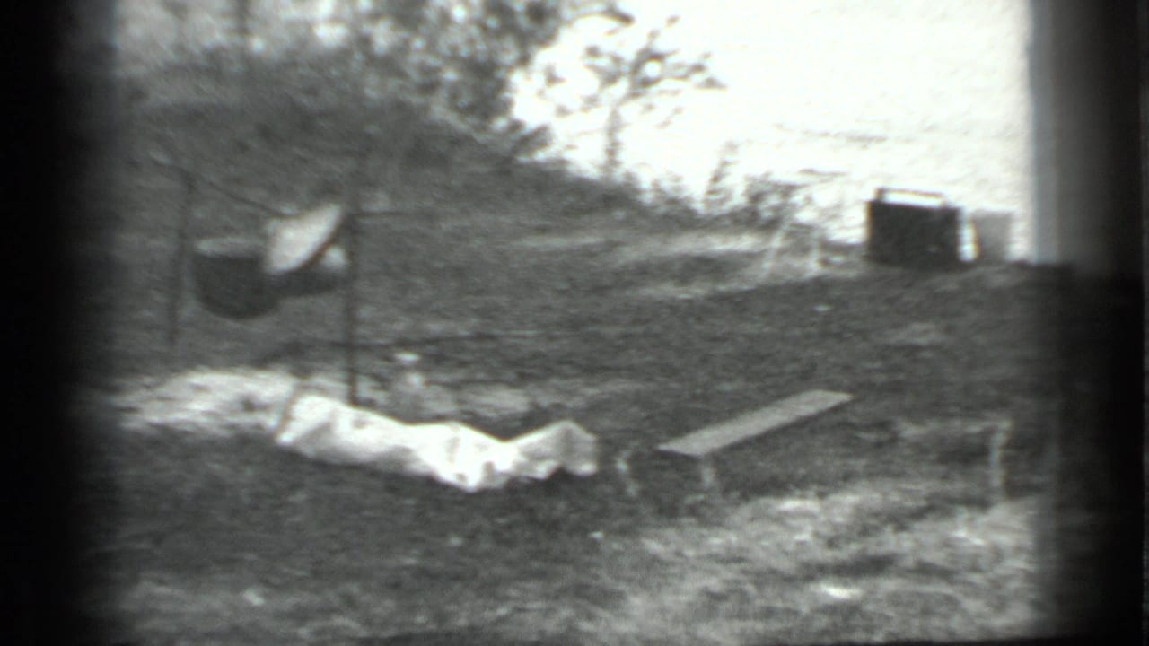Black-and-white Moscow 1983 campsite frame with a suspended pot, light tarp or cloth, a plank, brush, and a dark distant container or structure.