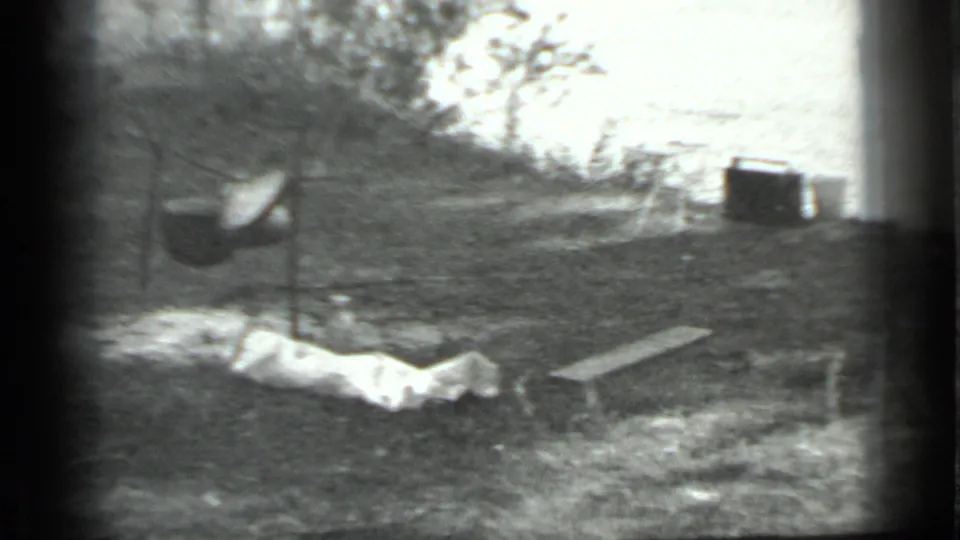 Black-and-white Moscow 1983 campsite frame with a suspended pot, light tarp or cloth, a plank, brush, and a dark distant container or structure.