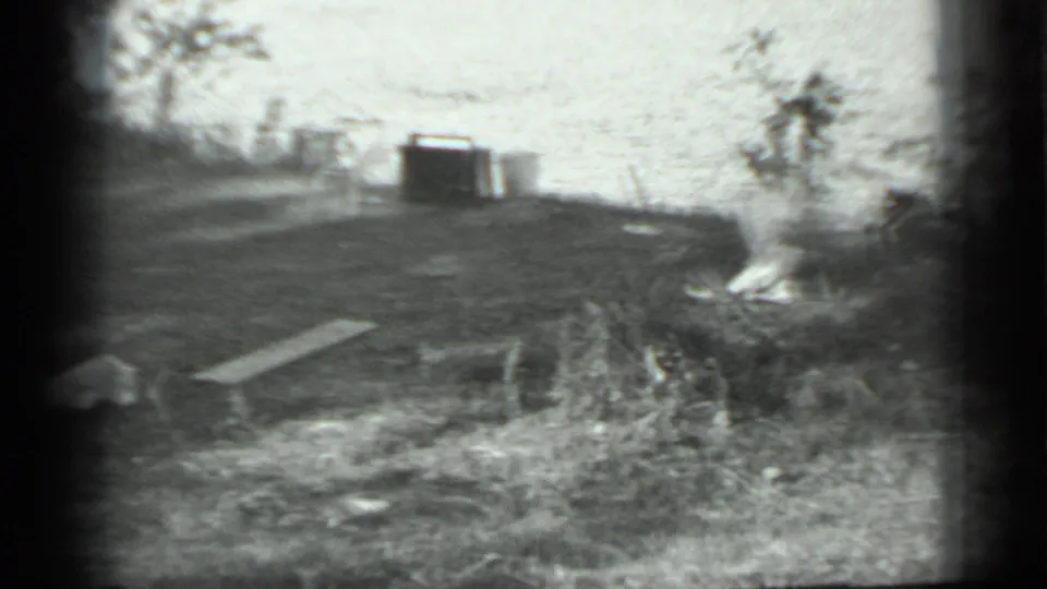 Black-and-white home-movie frame near Moscow showing a rough campsite area with a plank, brush, and a dark container or small structure beyond.