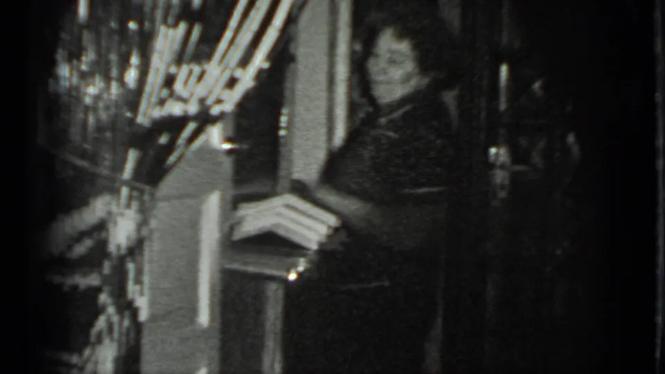In a grainy 1983 Moscow home-movie frame, an adult woman stands in a doorway near shelves and household objects.