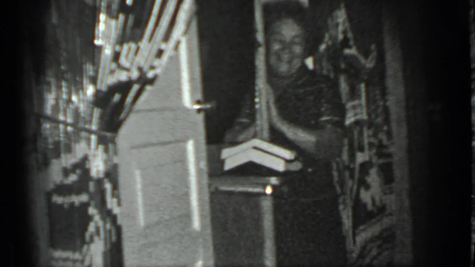 The woman remains in the doorway of a private Moscow interior, smiling within a soft, grainy 8mm home-movie frame.