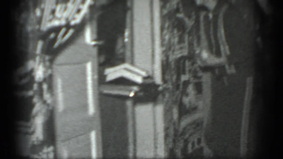 Grainy black-and-white Moscow home-movie frame showing a private interior with shelves, hanging items and a partial figure.
