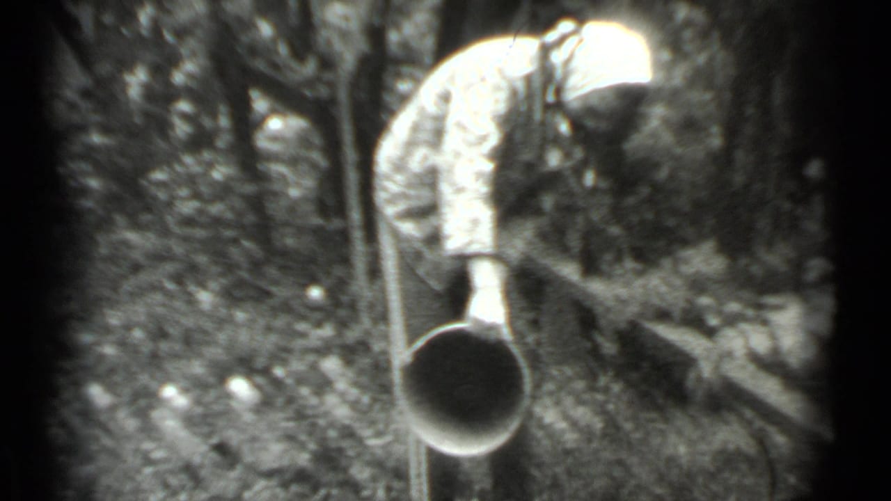 Woman in headscarf bends forward with a metal bucket among trees and bushes.