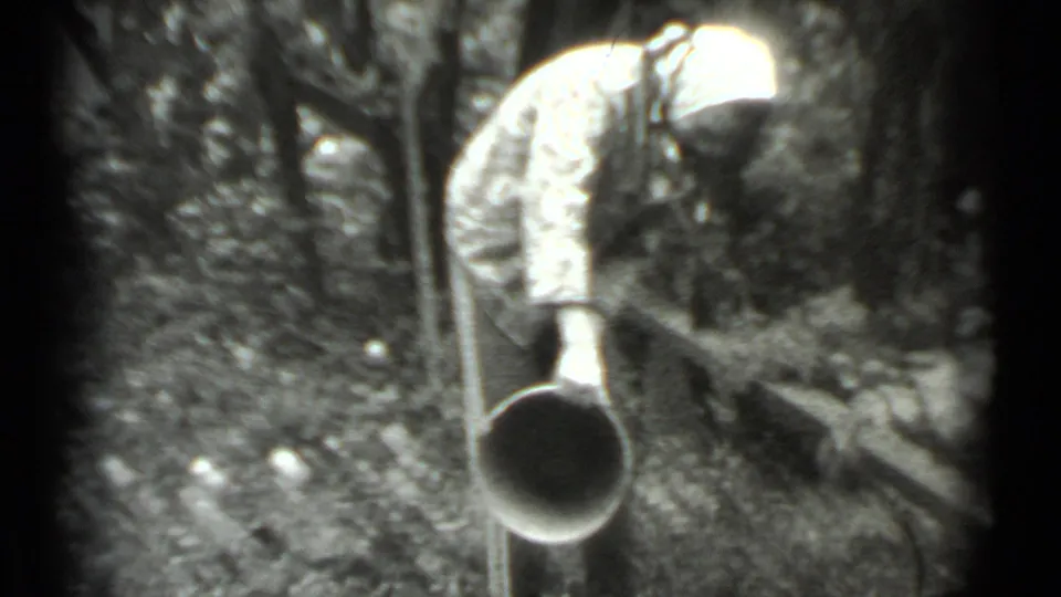 In supported 1983 Moscow home-movie footage, a woman in a headscarf bends with a metal bucket in a wooded area.