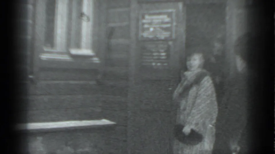 A woman in a patterned coat stands near a wooden doorway in the same 1983 Moscow home-movie scene.