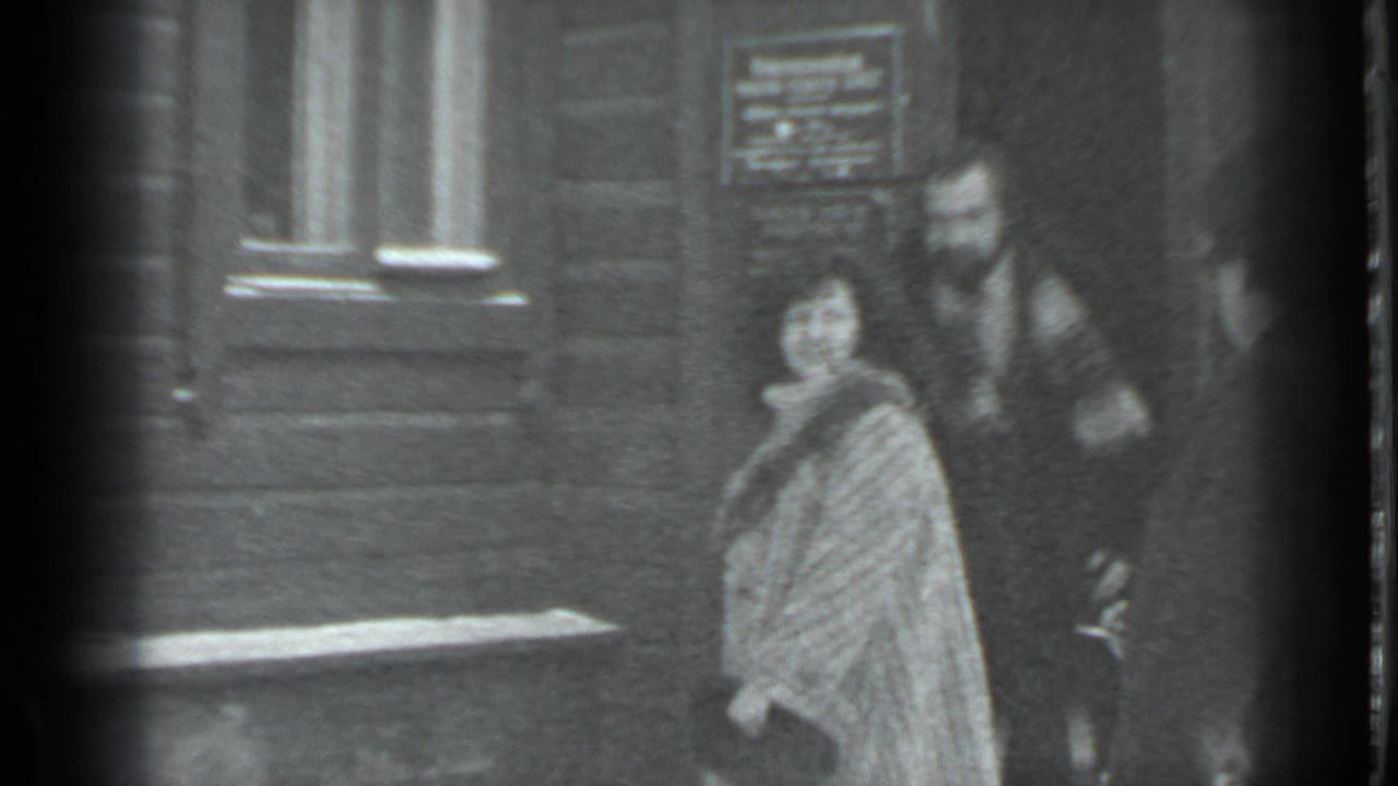 Woman in a patterned coat stands near a man outside a wooden building in snow.