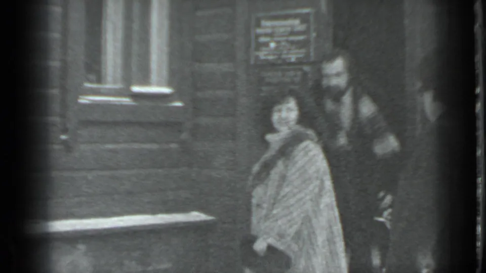 In 1983 Moscow home-movie footage, a woman in a patterned coat and a man in a dark coat stand beside a snowy wooden building.