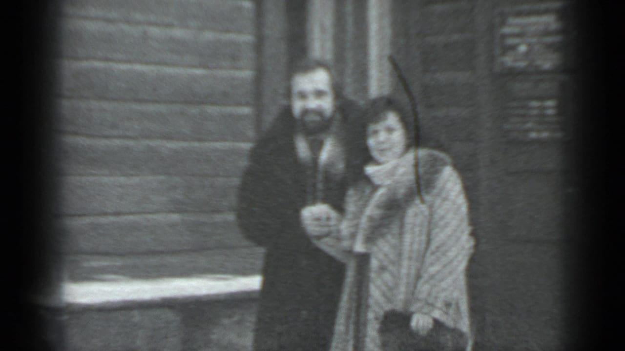 Man and woman stand close together by a wooden wall with snow at their feet.