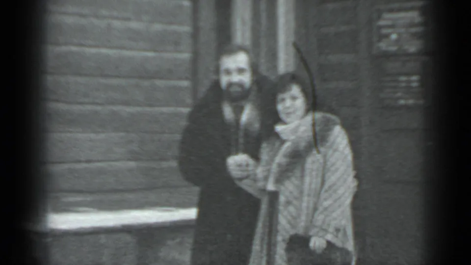 The couple faces the camera in Moscow, with the man in a dark coat and the woman in a patterned winter coat near a wooden exterior wall.