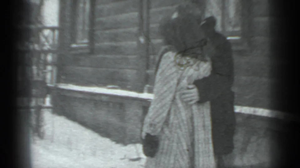 The man embraces the woman from behind as they stand beside a snowy wooden building in the 1983 Moscow home-movie clip.
