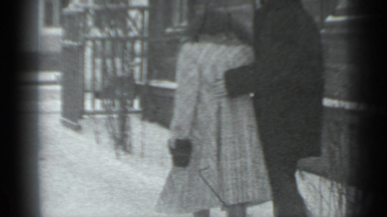 Two people in winter coats stand on a snowy street near a wooden exterior.