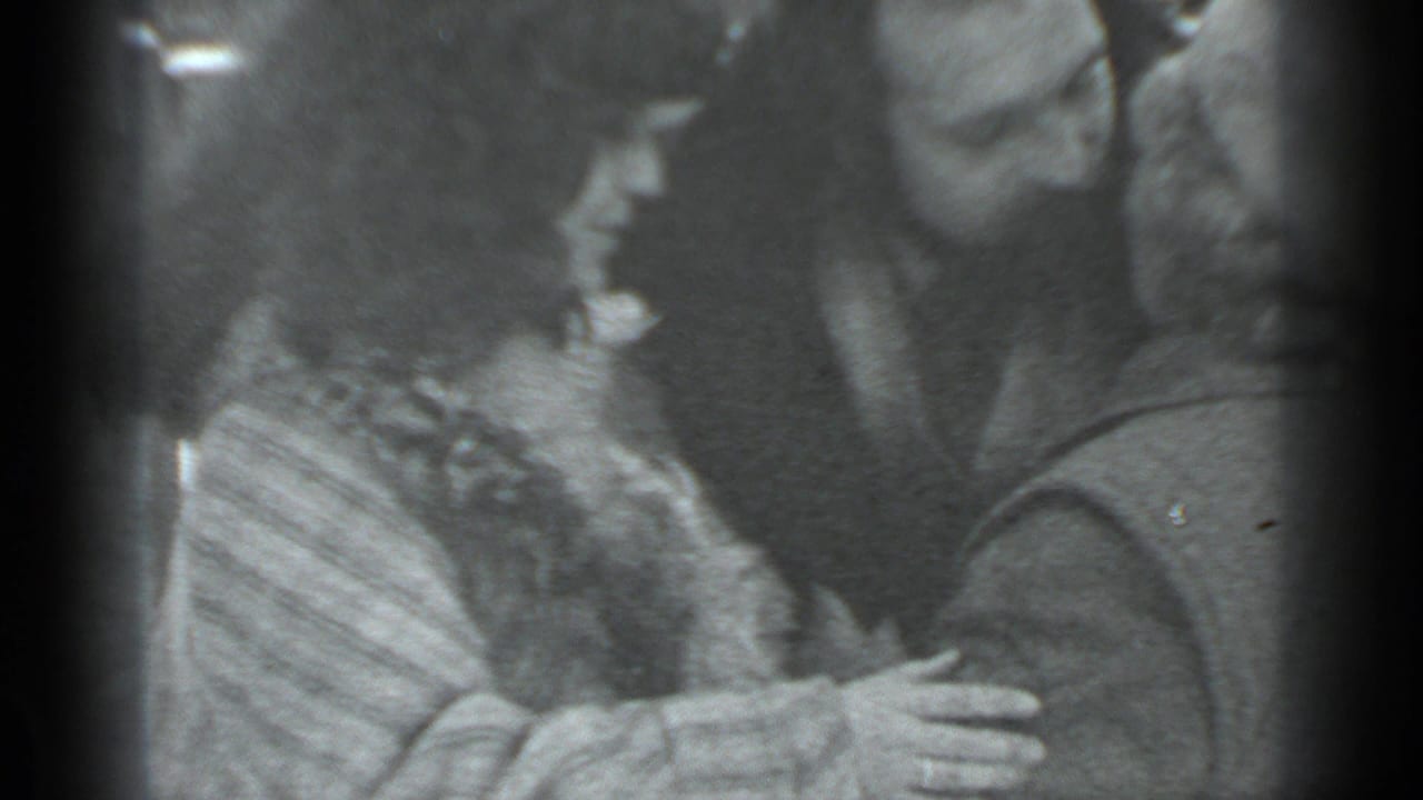 In the supplied 1983 Moscow home-movie context, three adults stand close together as a woman reaches toward a man during an affectionate greeting.