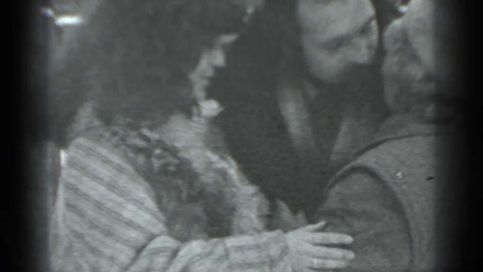 In the supplied 1983 Moscow home-movie context, three adults stand close together as a woman reaches toward a man during an affectionate greeting.