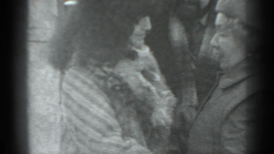A close black-and-white 8mm frame shows a woman and man facing each other closely, with another adult partly visible in the center.