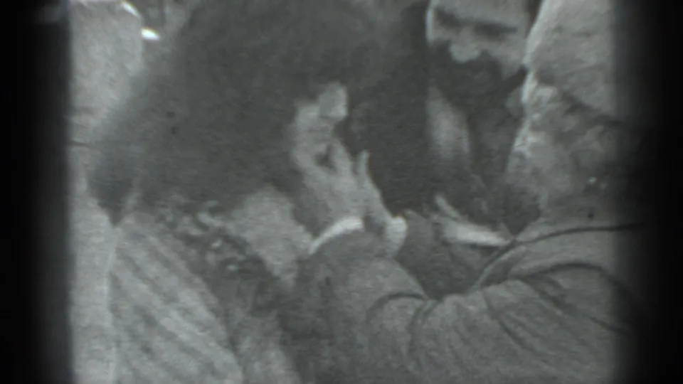 In this 8mm home-movie frame, two adults lean into a hug or kiss while another adult remains partly visible beside them.
