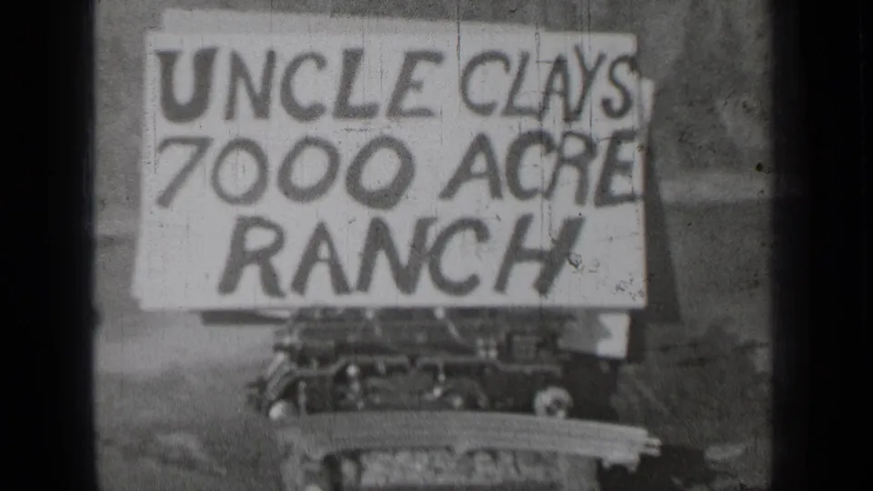Black-and-white 8mm frame of the Uncle Clays 7000 Acre Ranch sign, with soft focus and a vehicle or machine edge beneath it.