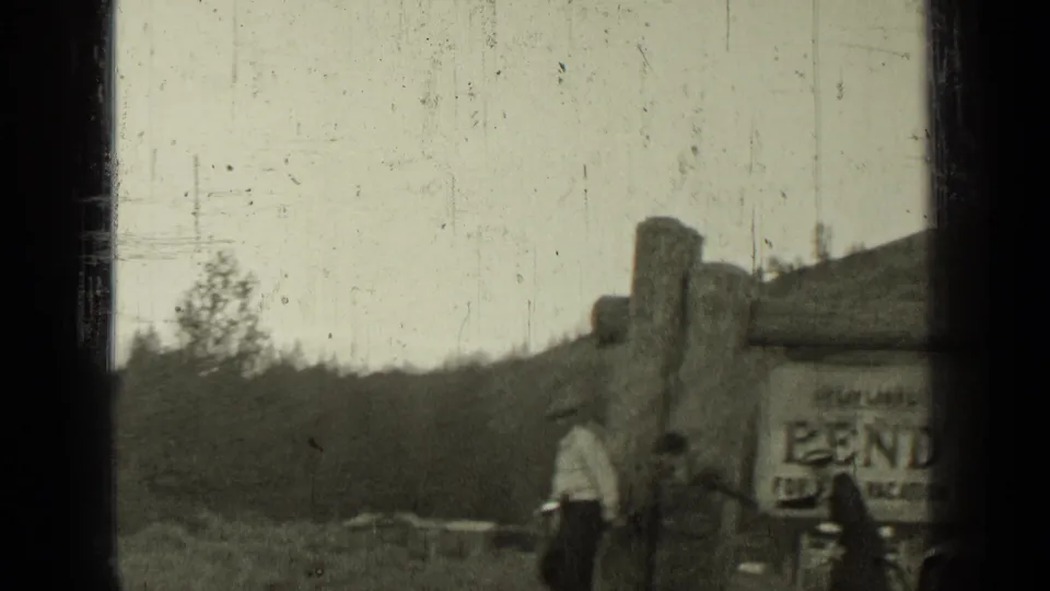 1937 Bend, Oregon frame showing people near a rustic roadside building with a Bend sign and wooded hills behind.