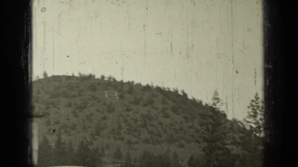 1937 Bend, Oregon home-movie frame with a wooded hillside behind the edge of a roadside structure.