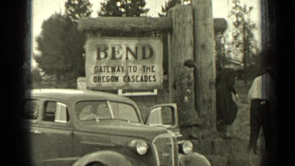 A 1930s car sits below the Bend gateway sign as adults in period clothing stand near the log structure.