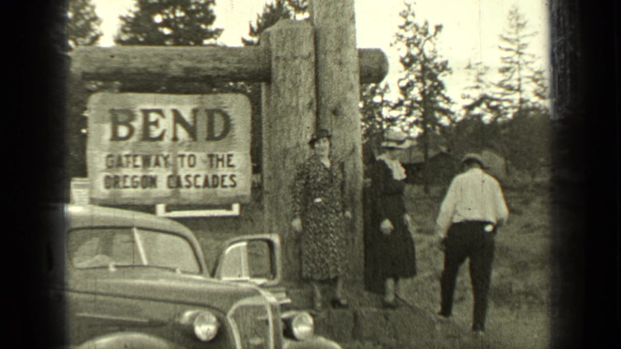 Visitors stand near a Bend sign, a parked car, and trees at a roadside stop.