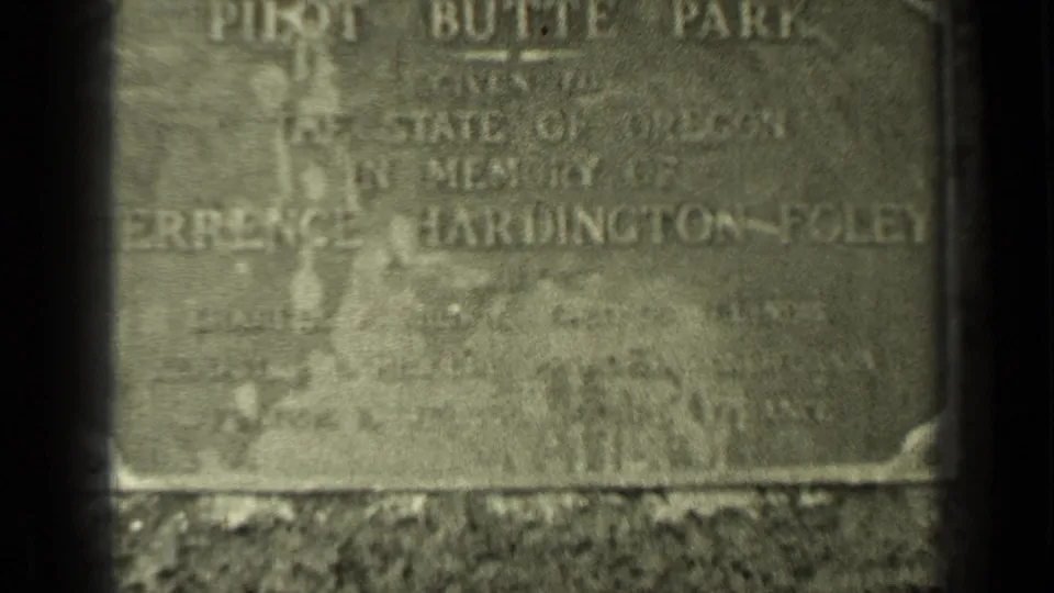 Close frame of the worn Pilot Butte Park marker, with engraved dedication text partly legible through soft 8mm focus and wear.