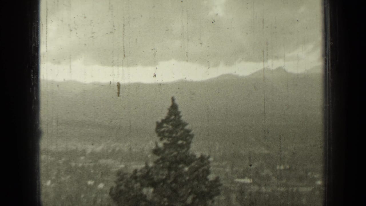 Window-framed view of cloudy mountains, a town below, and an evergreen tree in the foreground.