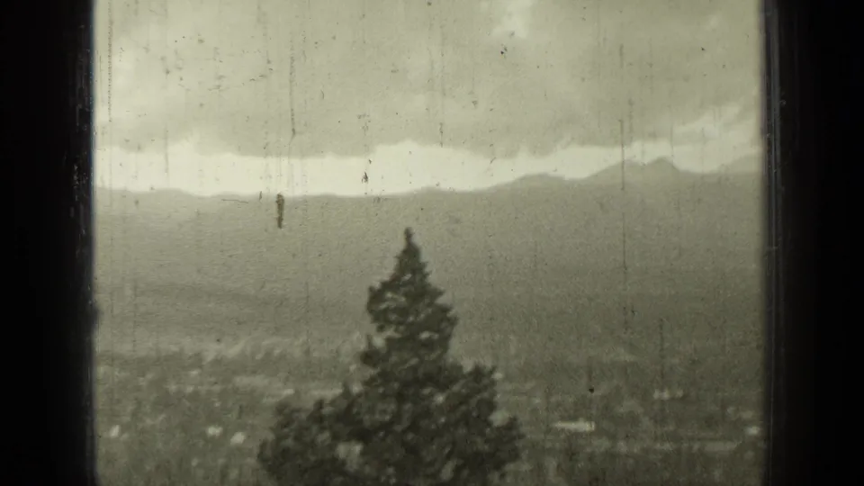 1937 Bend, Oregon home-movie frame showing distant mountains and town through a dark window frame, with an evergreen in front.