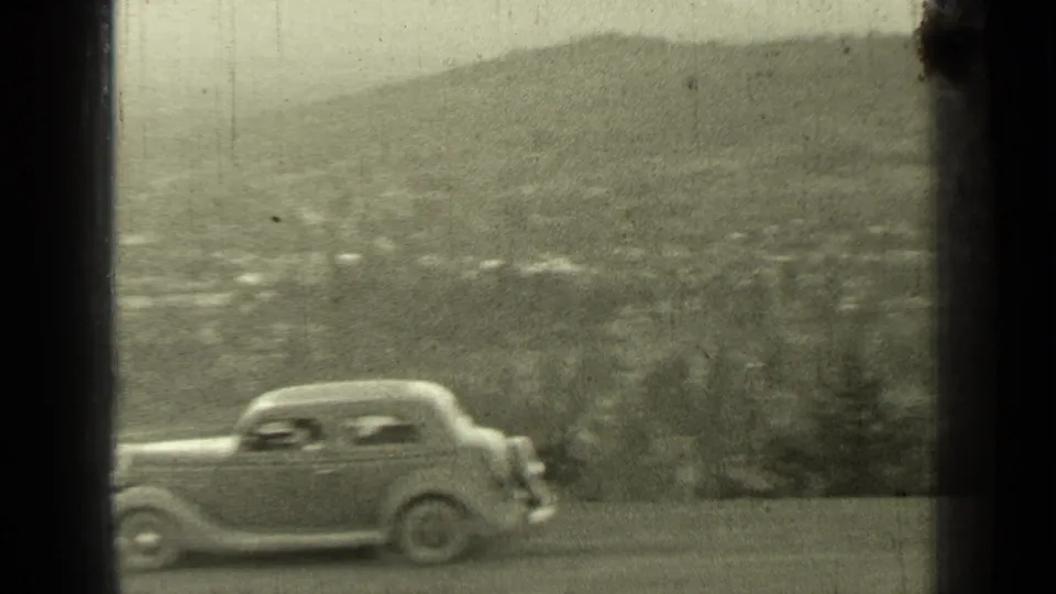 1937 Bend, Oregon frame with a light-colored period automobile passing in front of a distant hillside and town view.