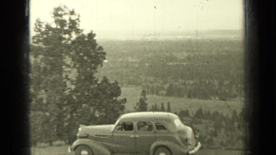 1937 Bend, Oregon frame showing a parked sedan with possible occupants, pine trees, and a broad valley overlook.
