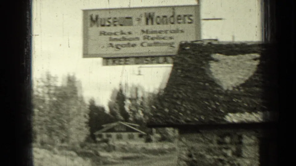 Wider 1937 Bend, Oregon frame showing the Museum of Wonders sign near a rustic building and tree-lined roadside setting.