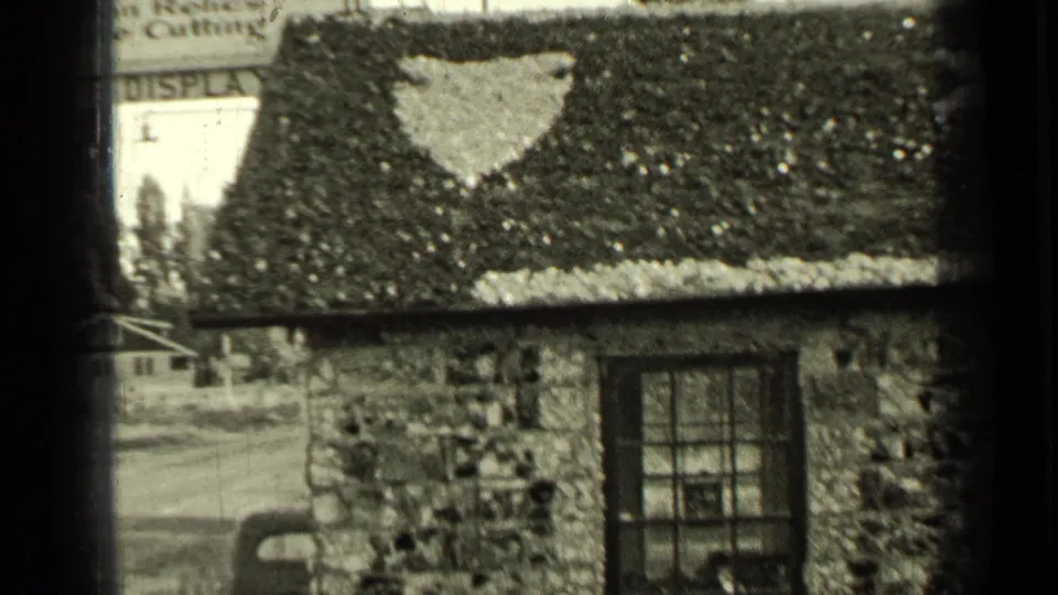 1937 Bend, Oregon home-movie frame showing a small stone building with a painted star-like roof shape, divided window, and partial sign in the background.