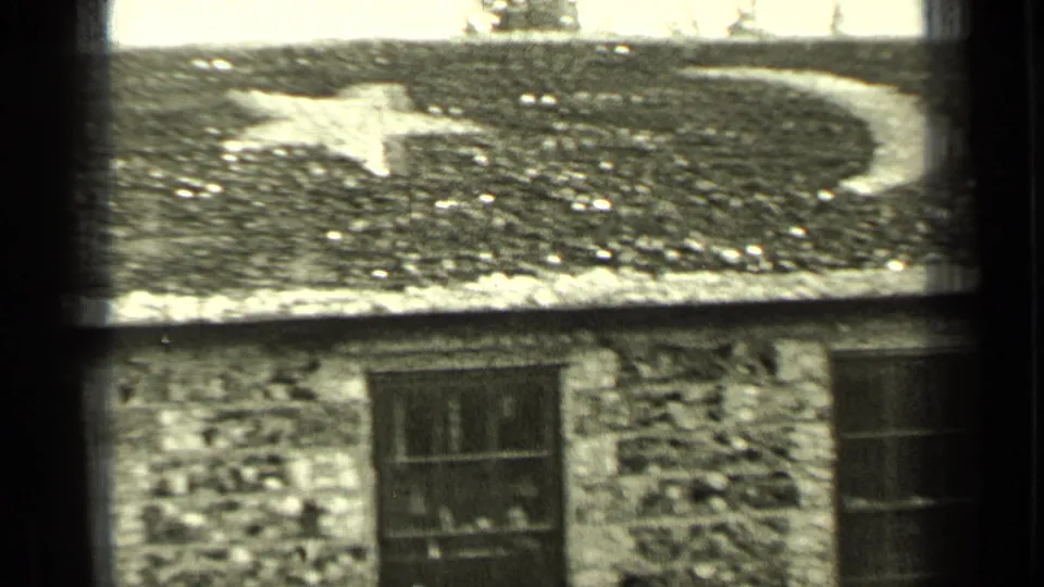1937 Bend, Oregon frame focusing on the decorated roof of a small stone building, with a pale moon-like shape above a dark divided window.
