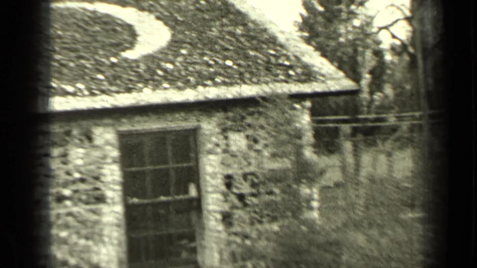 1937 Bend, Oregon home-movie view of the stone building from an angle, showing the moon-like roof decoration, window, fence, and trees.