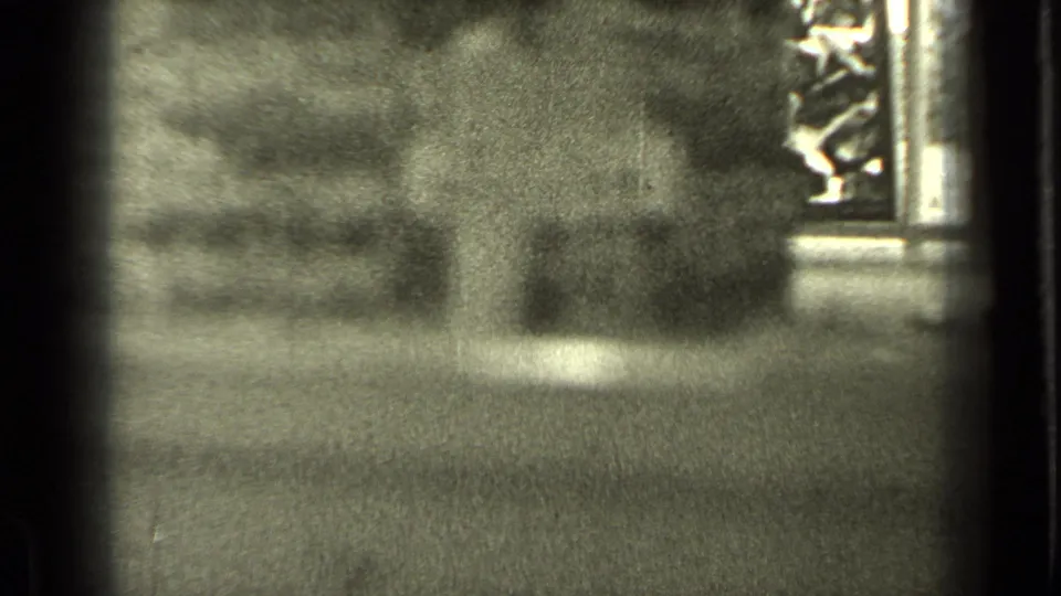 Grainy 1930s home-movie frame showing an unfocused window view with dark framing and outdoor shapes beyond.