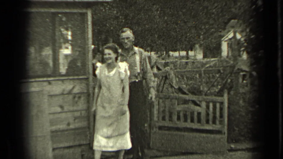 Young woman in a light dress and man in suspenders pose outside a wooden house in Bend, Oregon, 1937.