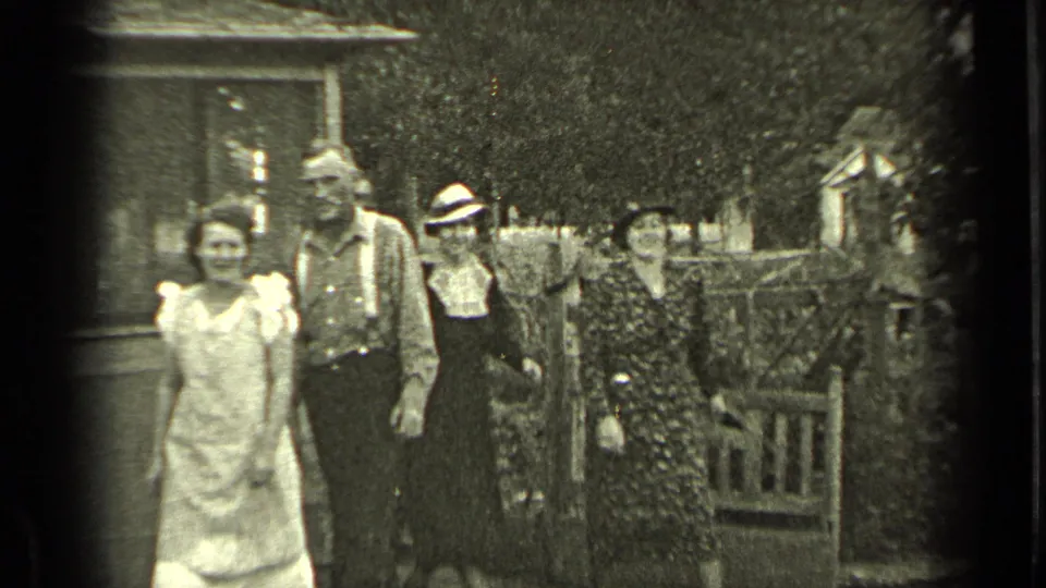 Group of adults in period dress stands outside a wooden house and garden gate in 1937 Bend, Oregon footage.