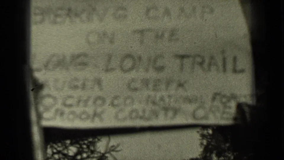 Close, soft-focus 8mm frame of a handwritten camp sign, apparently reading “Breaking Camp on the Long Trail,” with vegetation below.