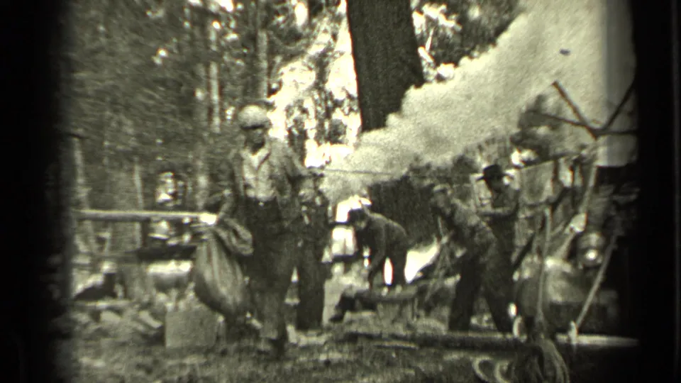 In Bend, Oregon, 1937, a man in work clothes carries a large sack past a smoky wooded campsite with adults and a dog nearby.